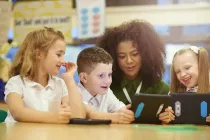 4 school children sat around a table smiling, chatting and looking at tablets together.