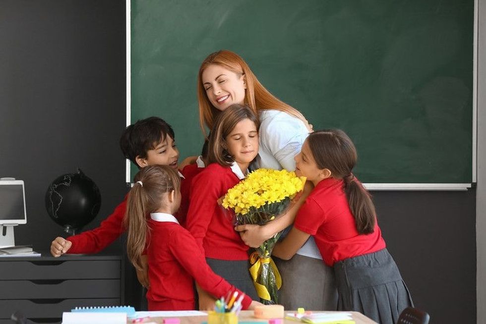 5 school children hugging their teacher in classroom with yellow flowers.