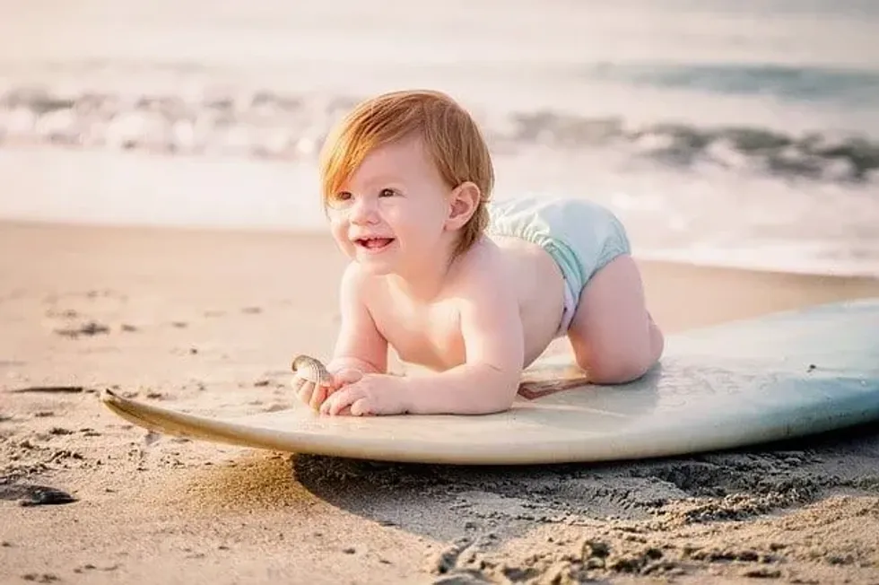A baby boy crawling on a surfboard on a beach.
