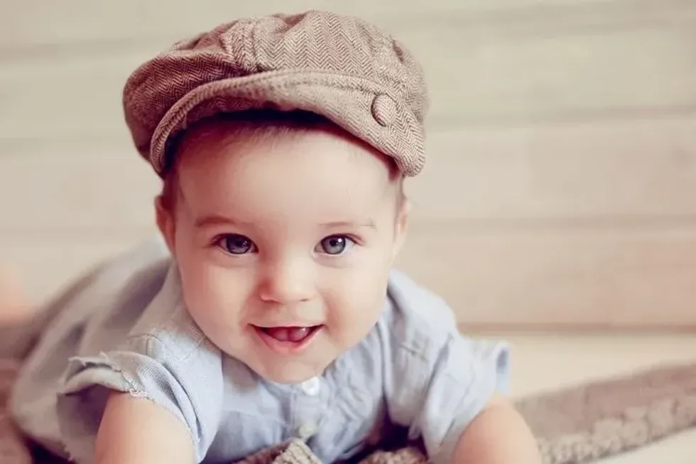 A baby boy wearing a brown hat and smiling looking at camera.