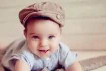 A baby boy wearing a brown hat and smiling looking at camera.