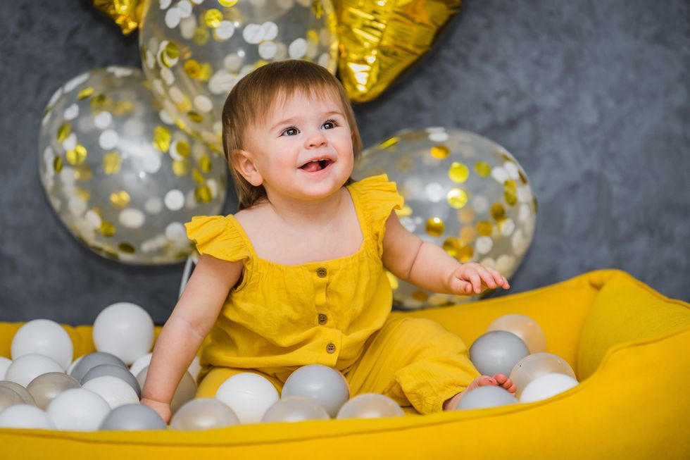 A baby girl in a yellow dress, gleeful among white balls, with gold and clear balloons in the background, echoing the charm of a melodic name for girls.
