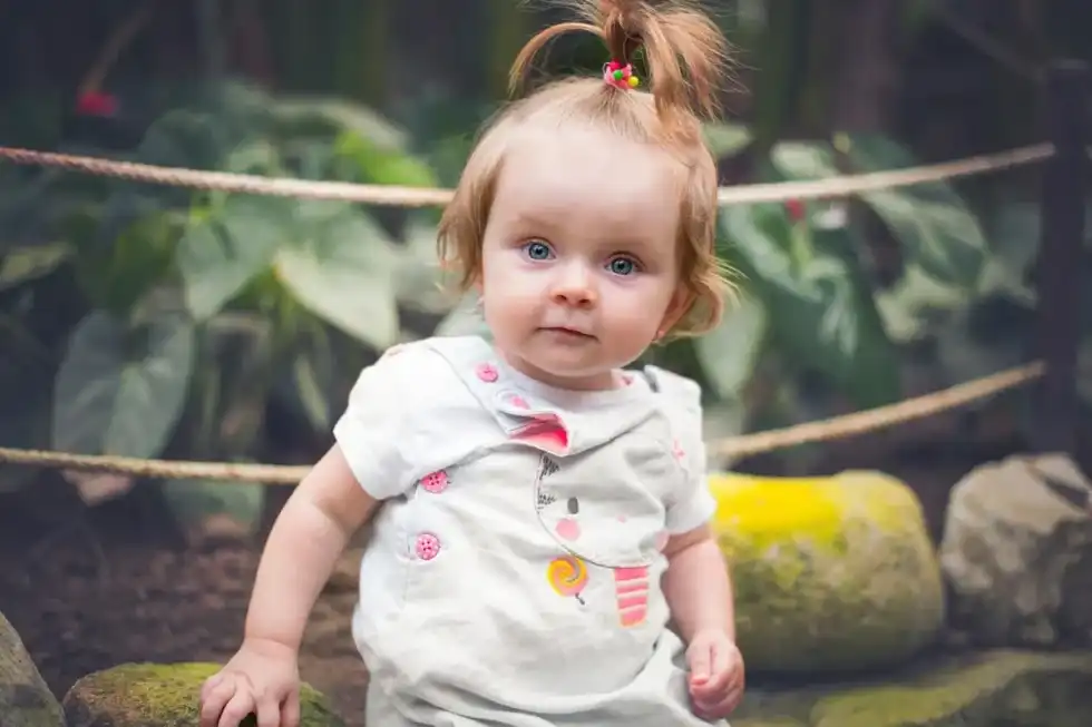 A baby girl with her hair in a cute ponytail stands and looks at the camera.