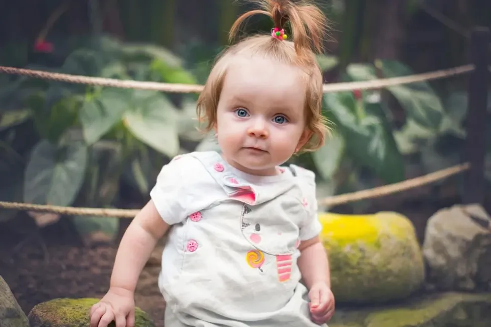A baby girl with her hair in a cute ponytail stands and looks at the camera.