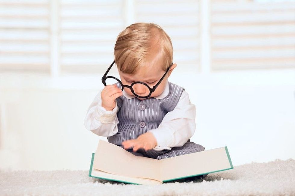 A baby holding a pair of glasses to him face with a book in front of him.