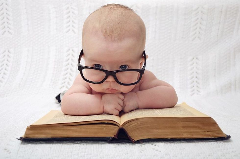 A baby wearing oversized glasses, leaning on a big book and staring ahead.