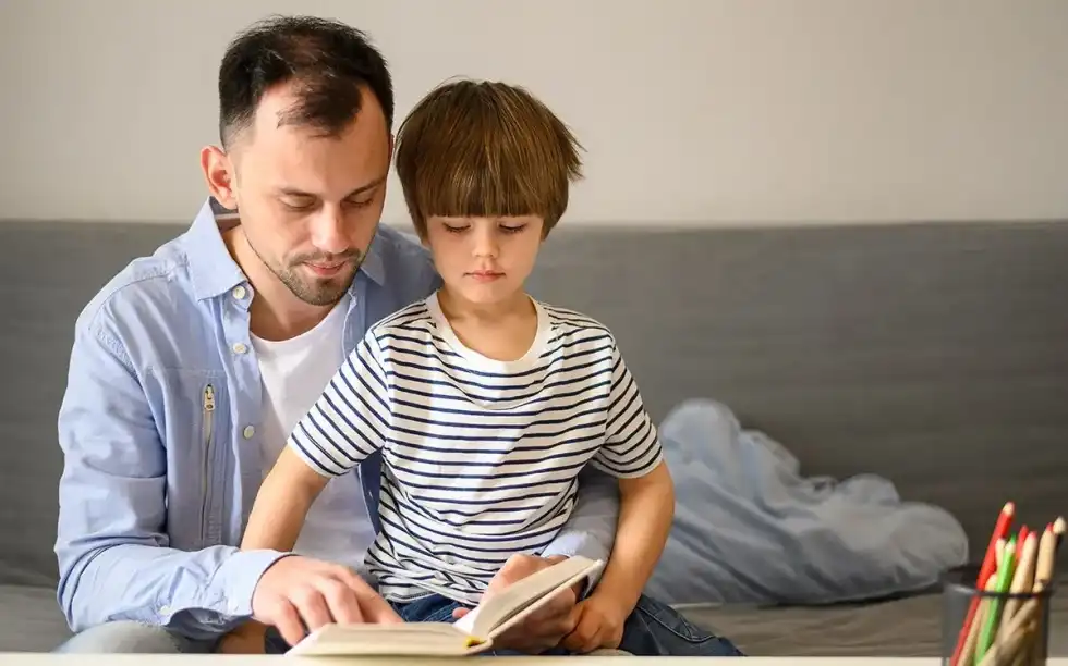 A boy sits on his father's lap as they read a book about WW2 art together.