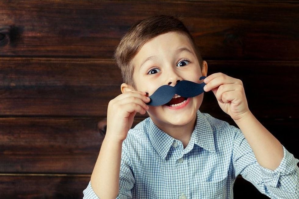 A cheerful boy holding a fake mustache to his face as if pondering over stupid riddles.