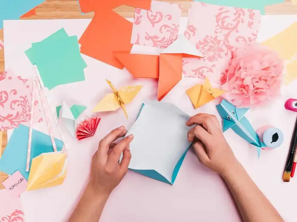 A child's hands surrounded by origami paper and models, making an origami basket.