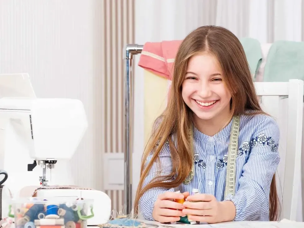 A girl using a sewing machine to make a DIY laptop case and surrounded by sewing equipment, including different coloured threads and a pin cushion, laughs into the camera.