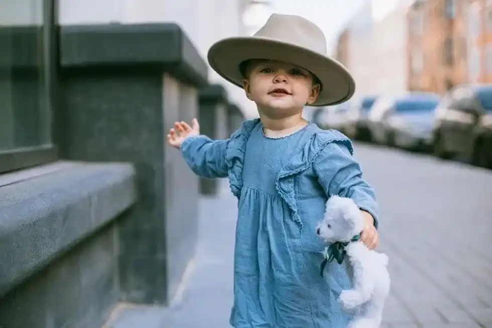 A littl egirl wearing blue dress and hat is holding her teddy bear