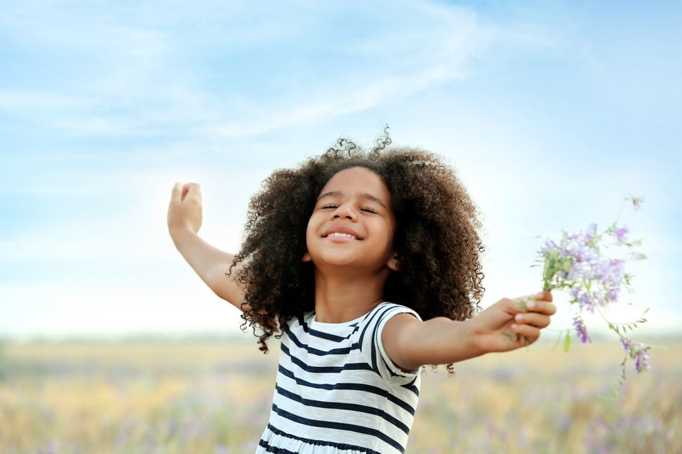 A little girl standing with arms outstretched in a grassy field, smiling contentedly, and holding out a bunch of flowers.