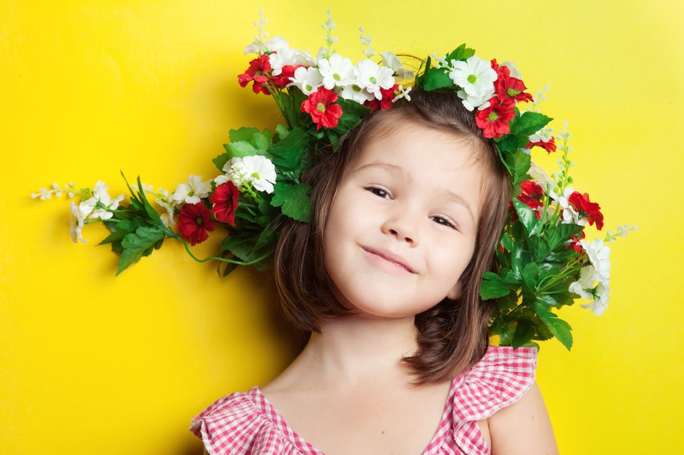 A little girl with a wreath of red and white flowers around her head, smiling and leaning against a yellow background.