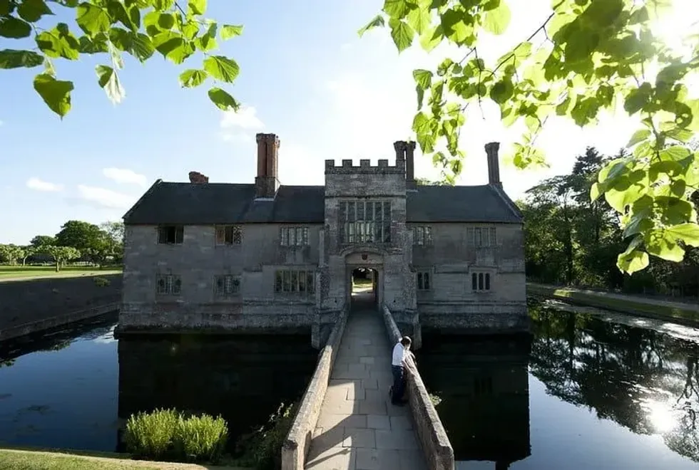 A moat, with two people standing on it looking at the water, leading up to a manor house.