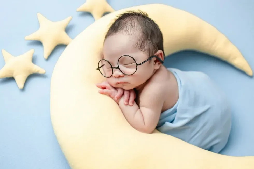 A newborn baby wearing glasses sleeping on moon pillow next to stars