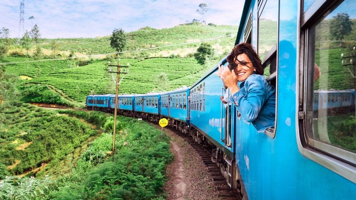 A passenger leans out of a bright blue train window, capturing the stunning tea plantation landscape as the train travels through a hilly, verdant region.