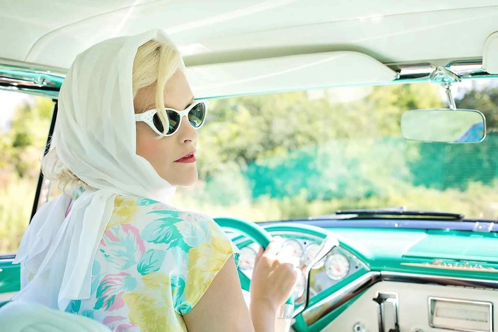 A retro woman wearing sunglasses and scarf driving a classic car in summer