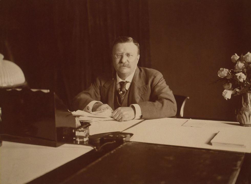 A sepia-toned photograph of President Theodore Roosevelt sitting at his desk with papers and a pen, looking directly at the camera.