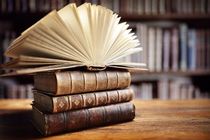 A stack of vintage leather-bound books on a wooden table, with one open displaying fanned pages, in a library setting for book trivia.