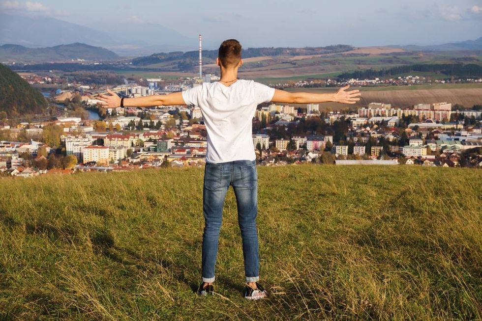 A view of a male standing on a hill with his arms open while proudly looking at his hometown.