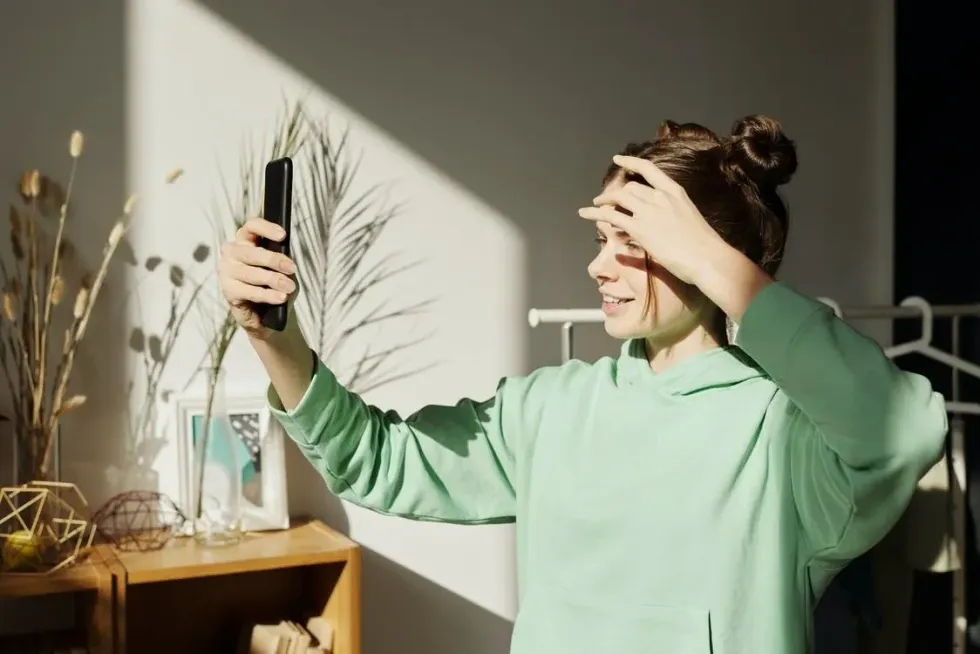 A woman in a mint green sweatshirt playfully covers her eyes with her hand, exuding a sense of confidence and self-acceptance.