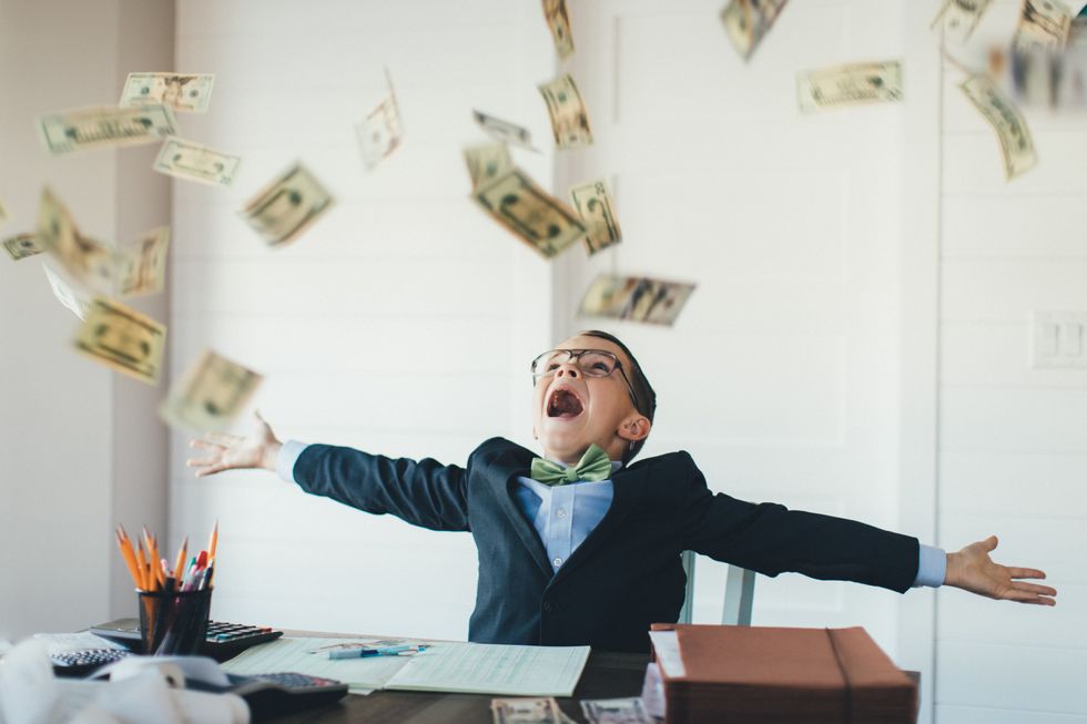 A young boy is dressed business attire with arms wide open and in excitement to catch US currency raining from the sky.