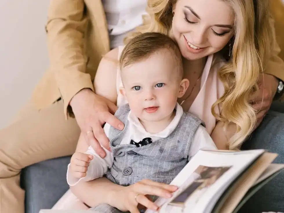 A young toddler sits on his mother's lap as his parents look through a baby scrapbook together.