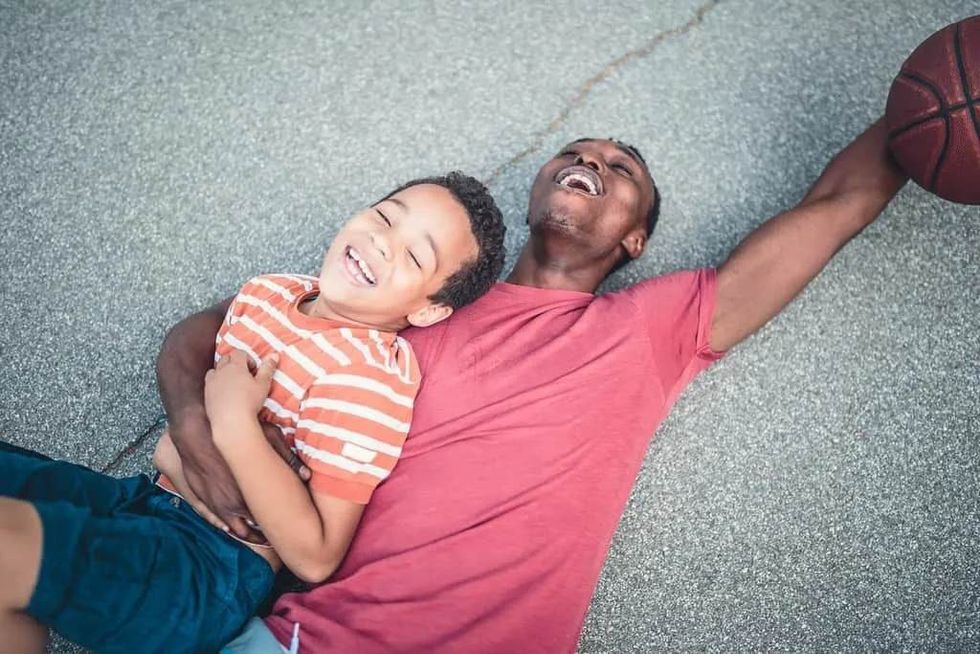 An adult and a child laughing on the floor of a basketball court, with the adul holding a basketball.