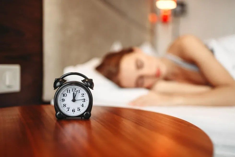 An alarm clock on a bedside table with a young woman asleep in rhe background