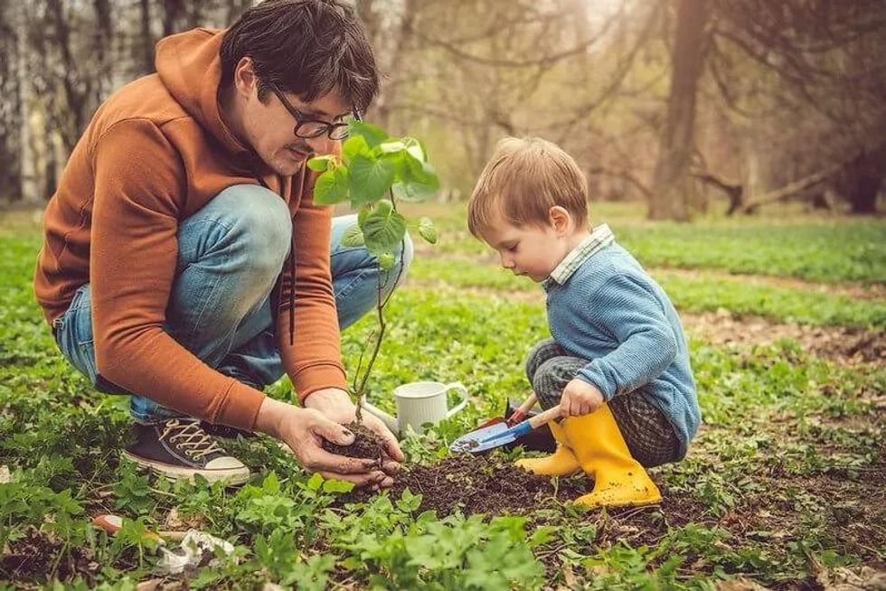 An image showing father and son squatting to garden. The son holding a mini shovel while the father has his handful with soil