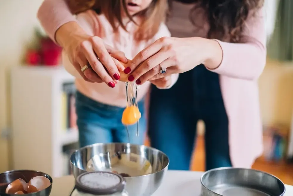 An Image showing the hands of a mother and daughter breaking eggs into a pan 