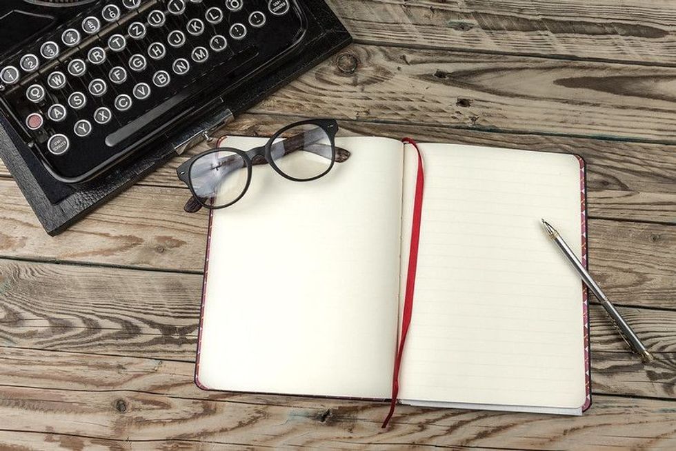 An open book with a red page stopper, a pen, and a pair of glasses on a wooden table, with a typewriter in the background