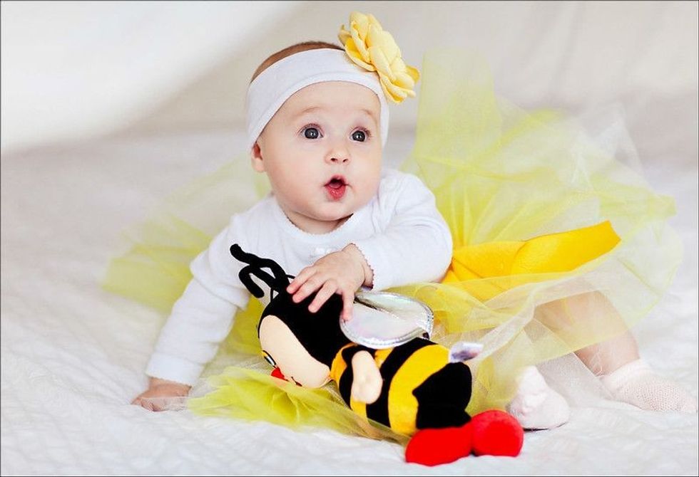 Baby girl wearing flower headband sitting on bed with bee toy