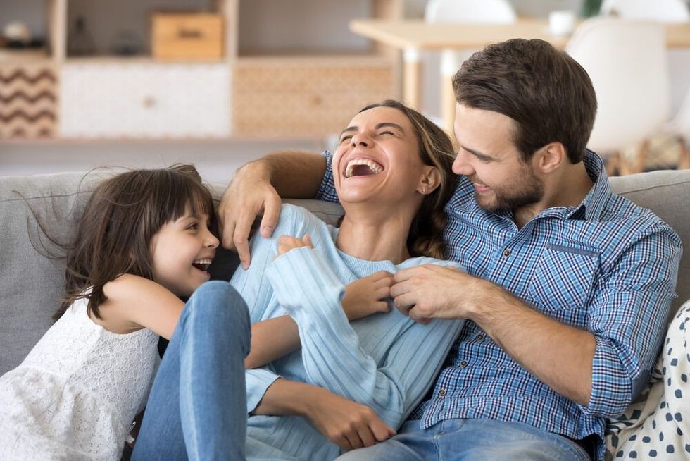 Cheerful people sitting on couch in living room have fun little daughter tickling mother laughing together with parents