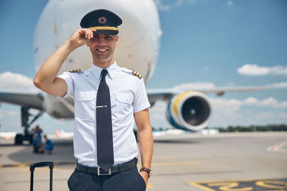 Cheerful young man airline worker touching captain hat and smiling while standing in airfield with airplane on background
