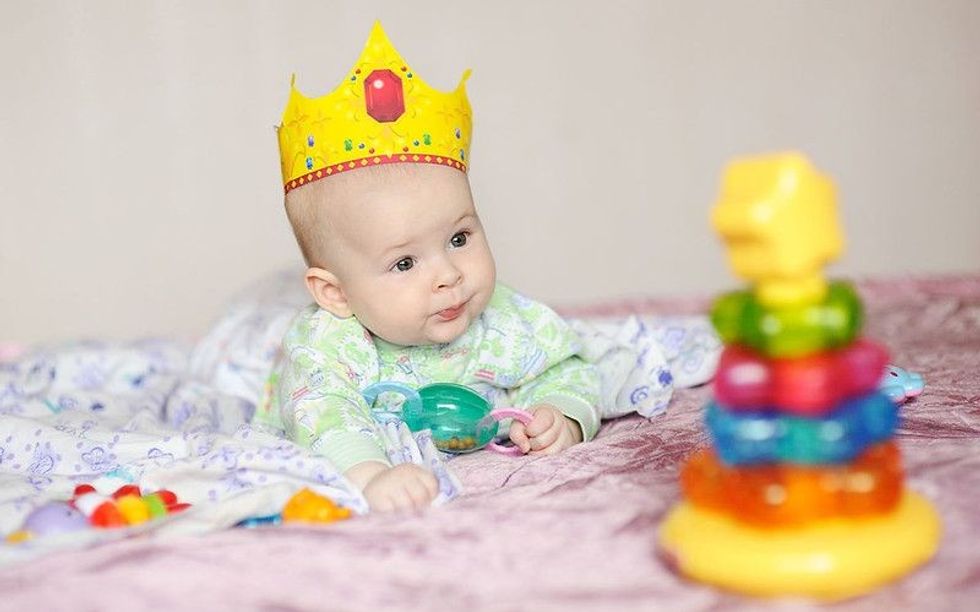 Child wearing crown lies on a bed with toys.
