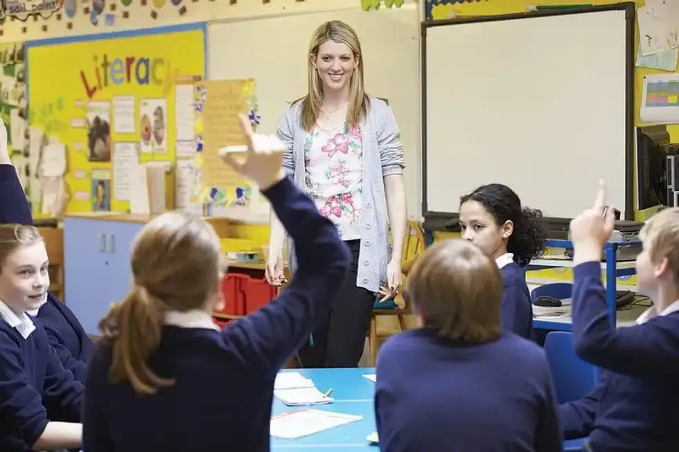 Children in a class room setting, with a teacher standing infront of them.