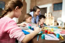 Children learning to paint with art materials on a surface and a teacher in the background.
