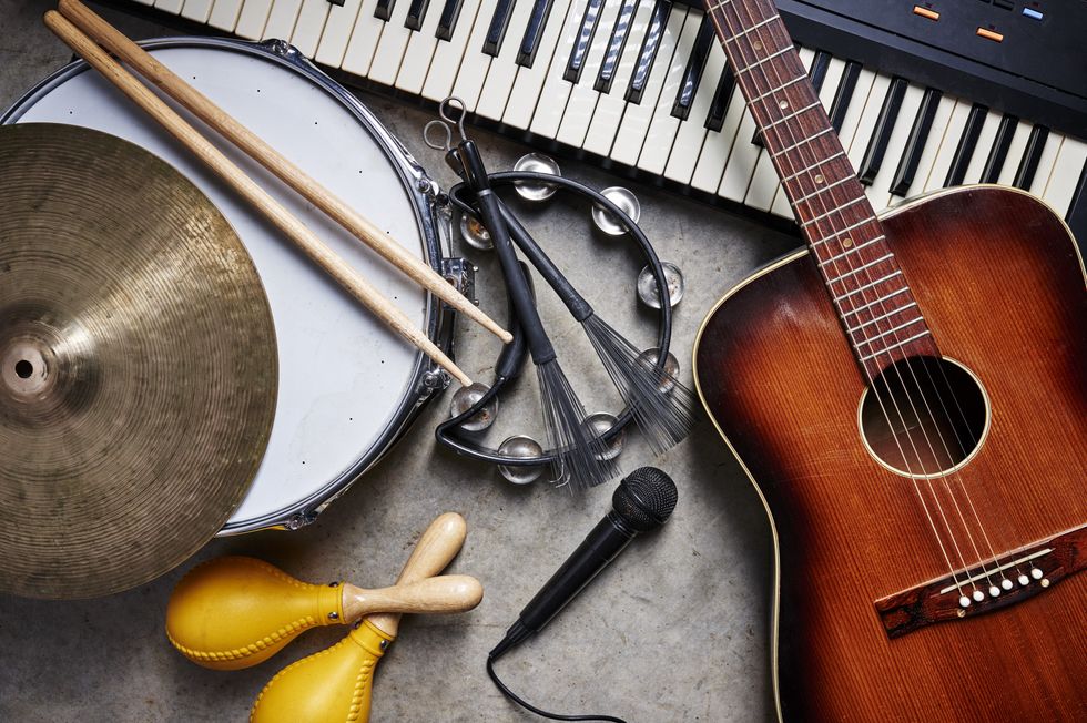 Collection of musical instruments including a guitar, keyboard, drum, cymbal, tambourine, maracas, and a microphone arranged on a concrete floor, ready for a jam session.