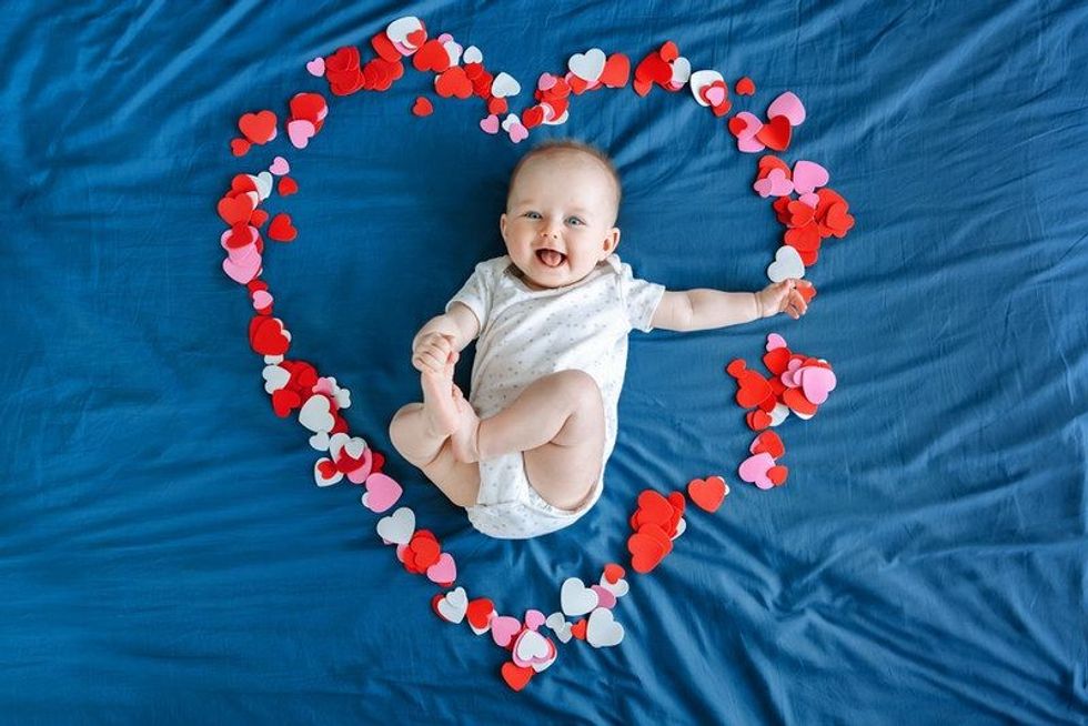 Cute smiling white Caucasian baby girl infant with blue eyes, four months old lying on bed in many foam paper and red pink colorful hearts.
