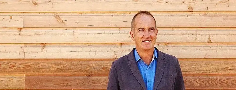 Designer Kevin McCloud standing in front of a wooden panelling smiling. 