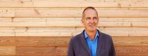 Designer Kevin McCloud standing in front of a wooden panelling smiling.