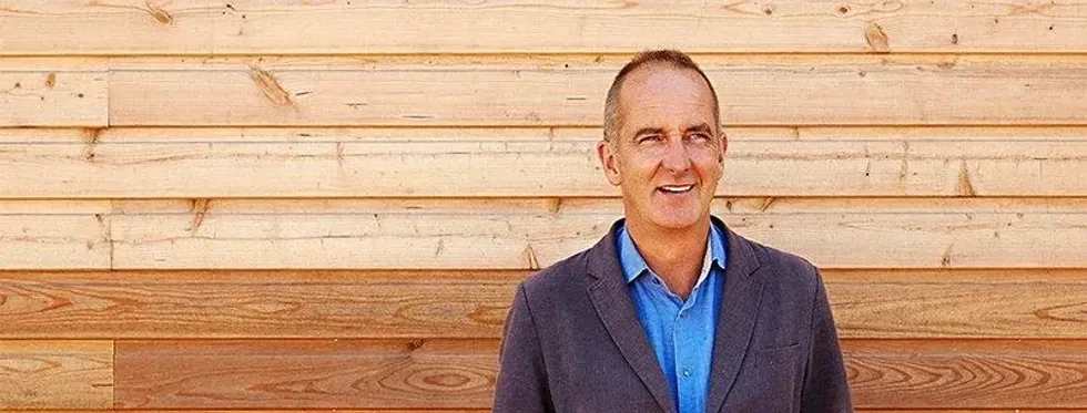 Designer Kevin McCloud standing in front of a wooden panelling smiling. 