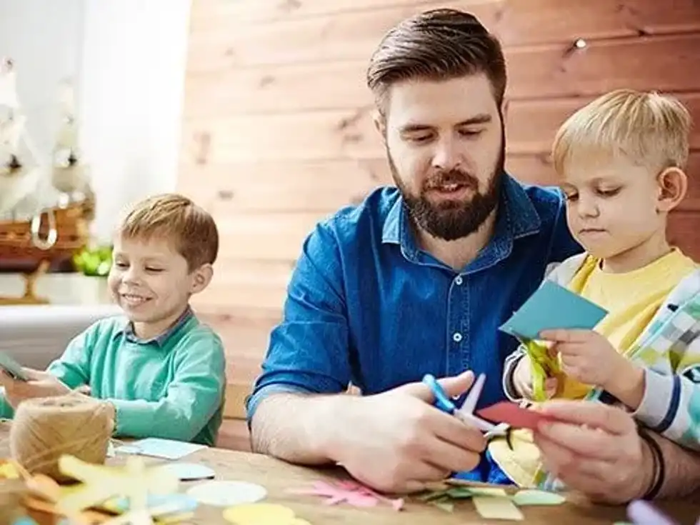 Father sat at the table with his sons, they are all cutting paper to make origami leaves.