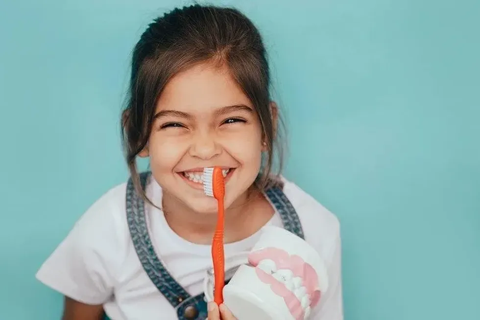 Girl holding a giant toothbrush and fake teeth grinning in front of a blue background.