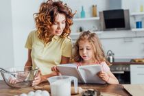 Girl reading cookbook near mother holding glass bowl with whisk.