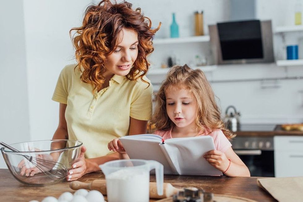 Girl reading cookbook near mother holding glass bowl with whisk