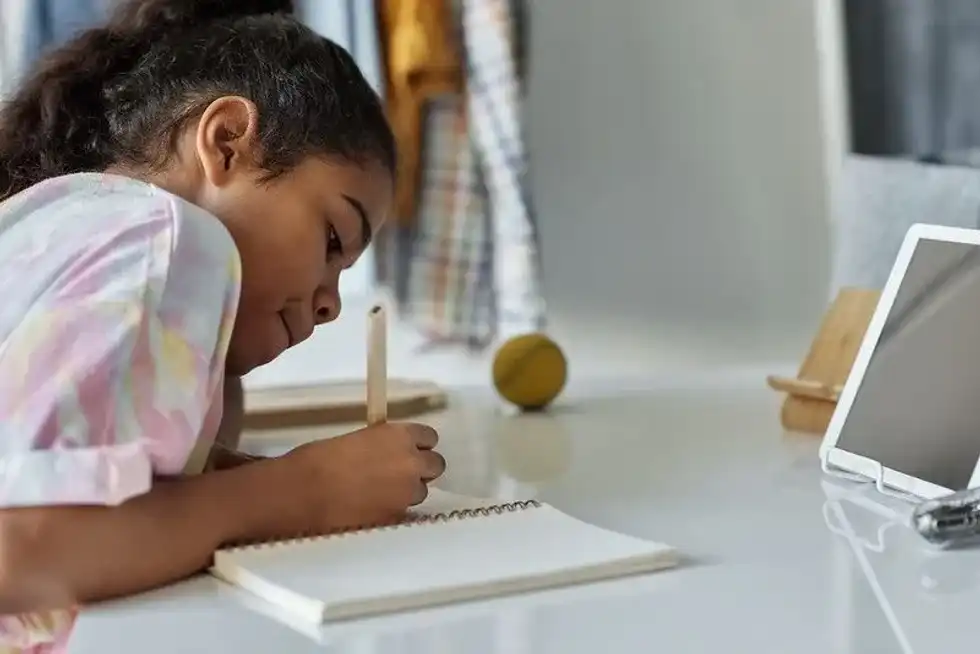 Girl sat at desk writing in a notebook and looking at a tablet.
