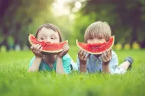 Happy boys lying on the grass on their tummies making a smile with a slice of watermelon.