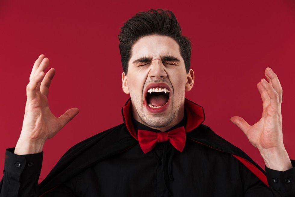 Image of angry vampire man with blood and fangs in black halloween costume screaming isolated over red wall